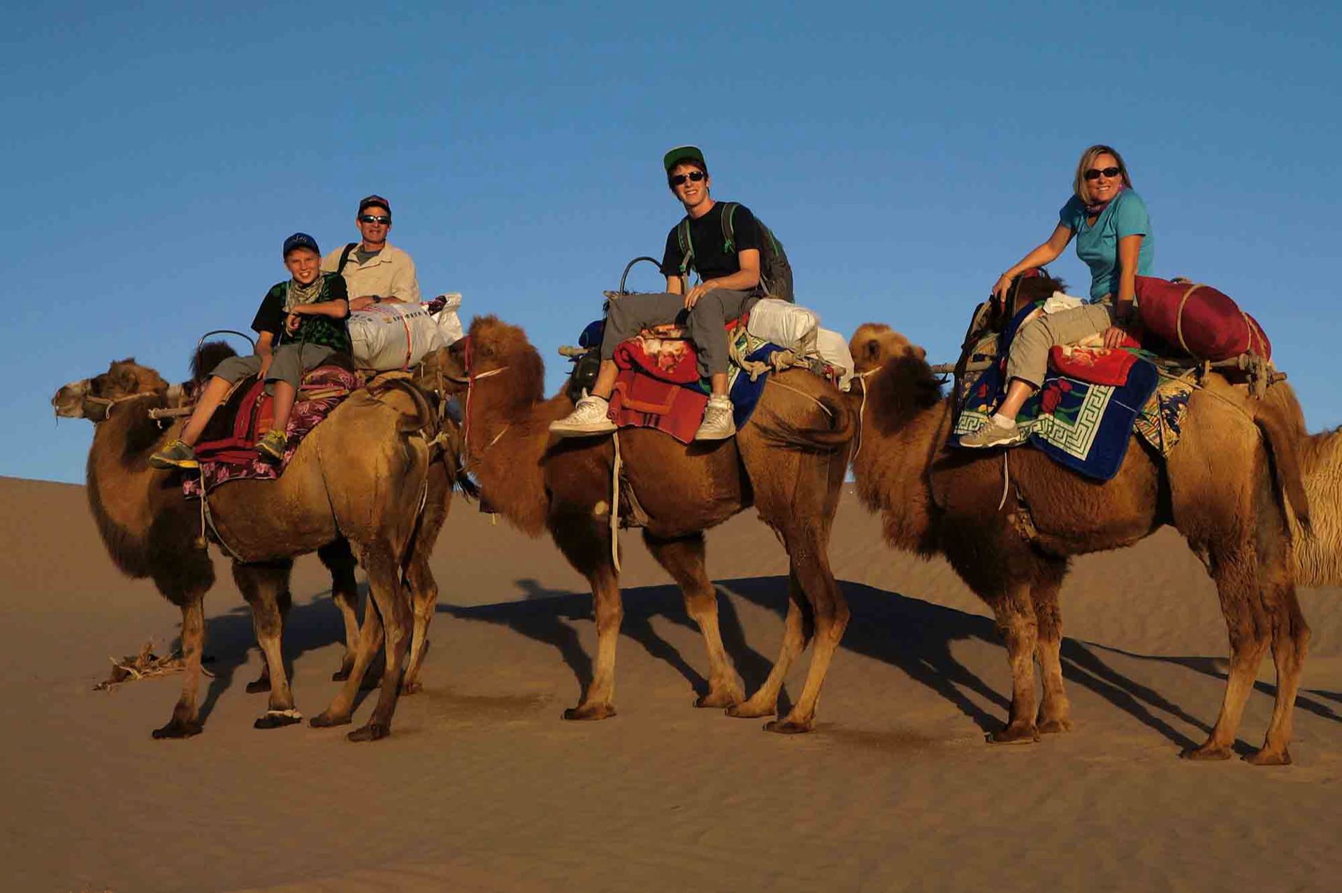 European travelers riding Bactrian camels across the Taklamakan Desert at golden hour — a Silk Road journey through Xinjiang, China.